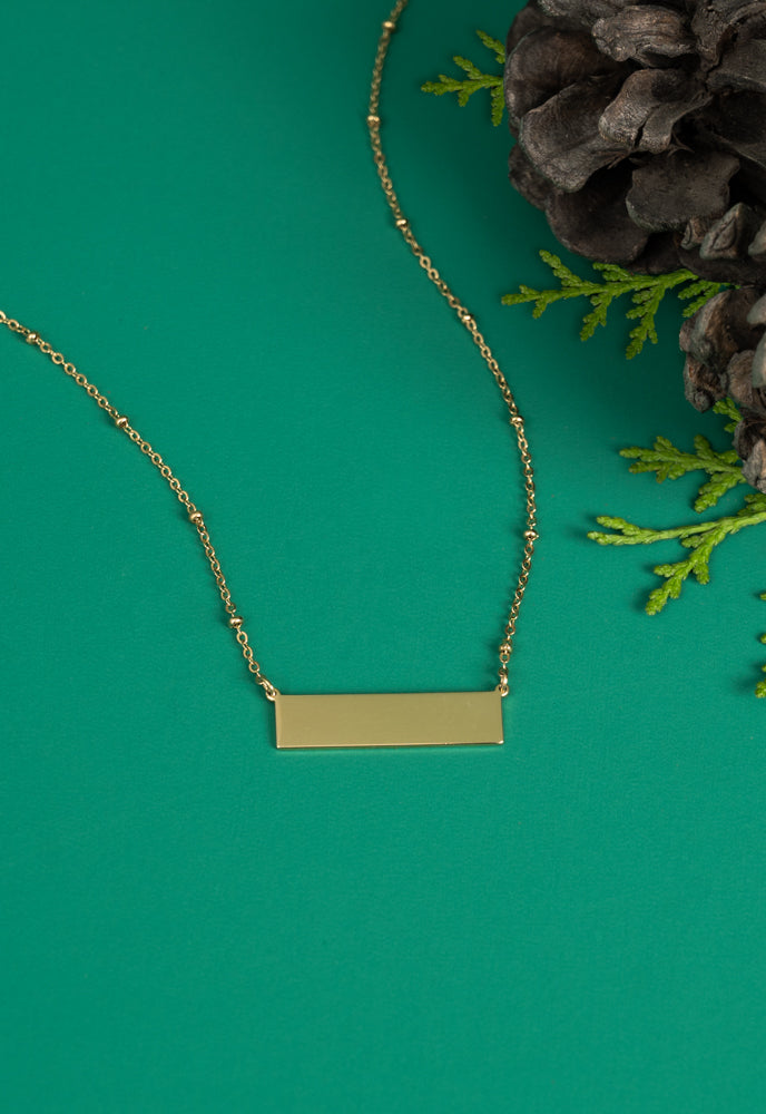 Engravable gold bar necklace on green background with pinecone and greenery