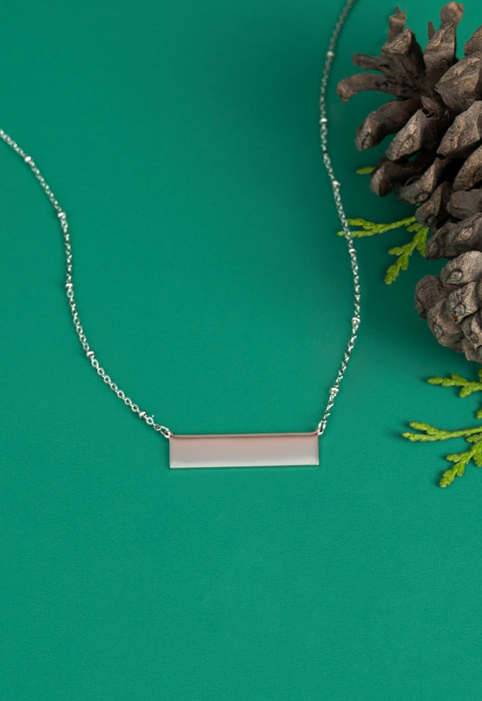 Engravable silver bar necklace on green background with pinecone and greenery