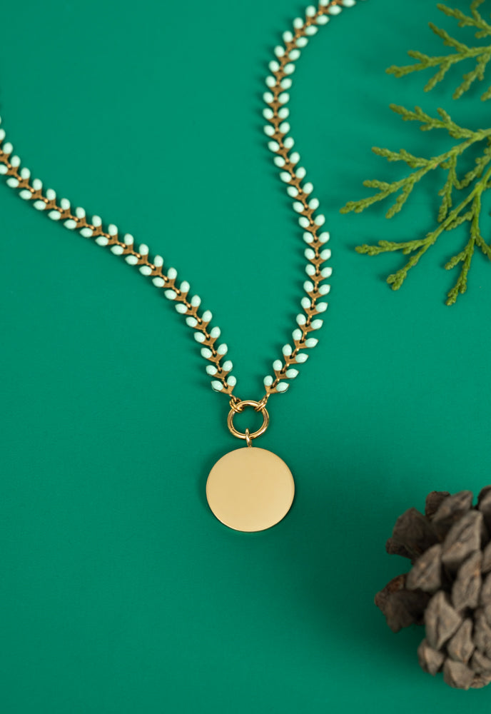 Engravable round pendant on mint and gold Seeds of Hope chain on green with evergreen sprigs