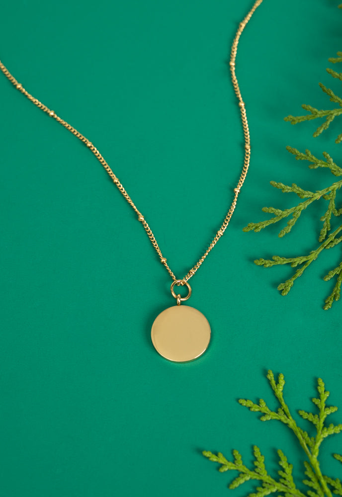 Engravable round gold pendant necklace on green with evergreen sprigs