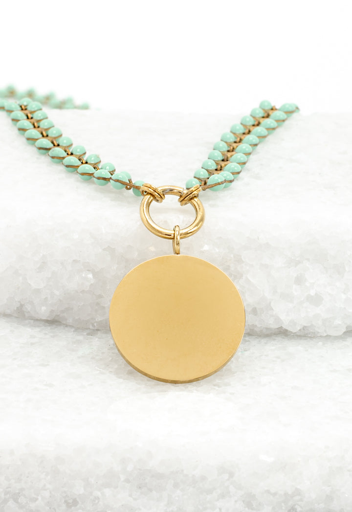 Engravable gold round pendant on mint enamel Seeds of Hope leaf chain resting on white stone