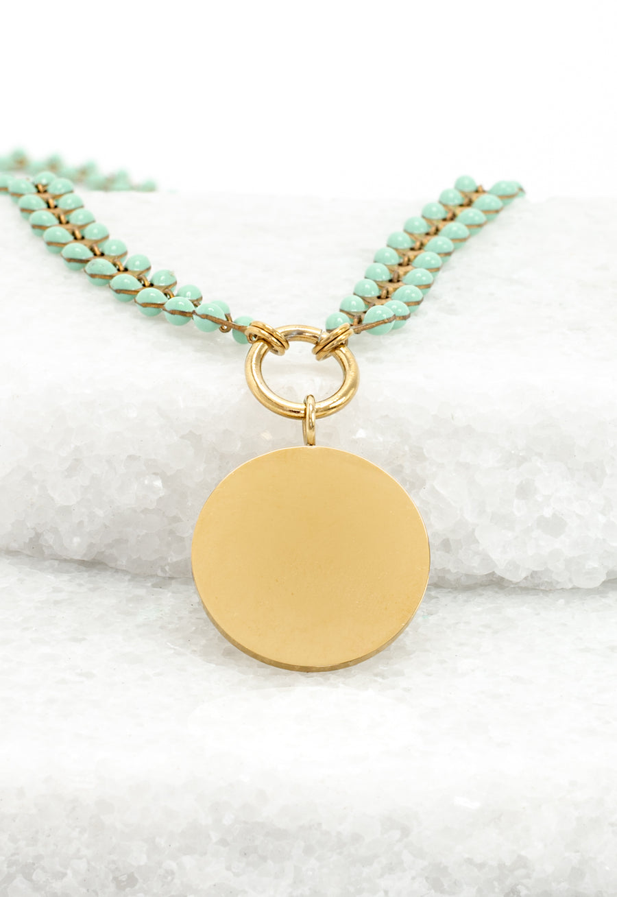 Engravable gold round pendant on mint enamel Seeds of Hope leaf chain resting on white stone